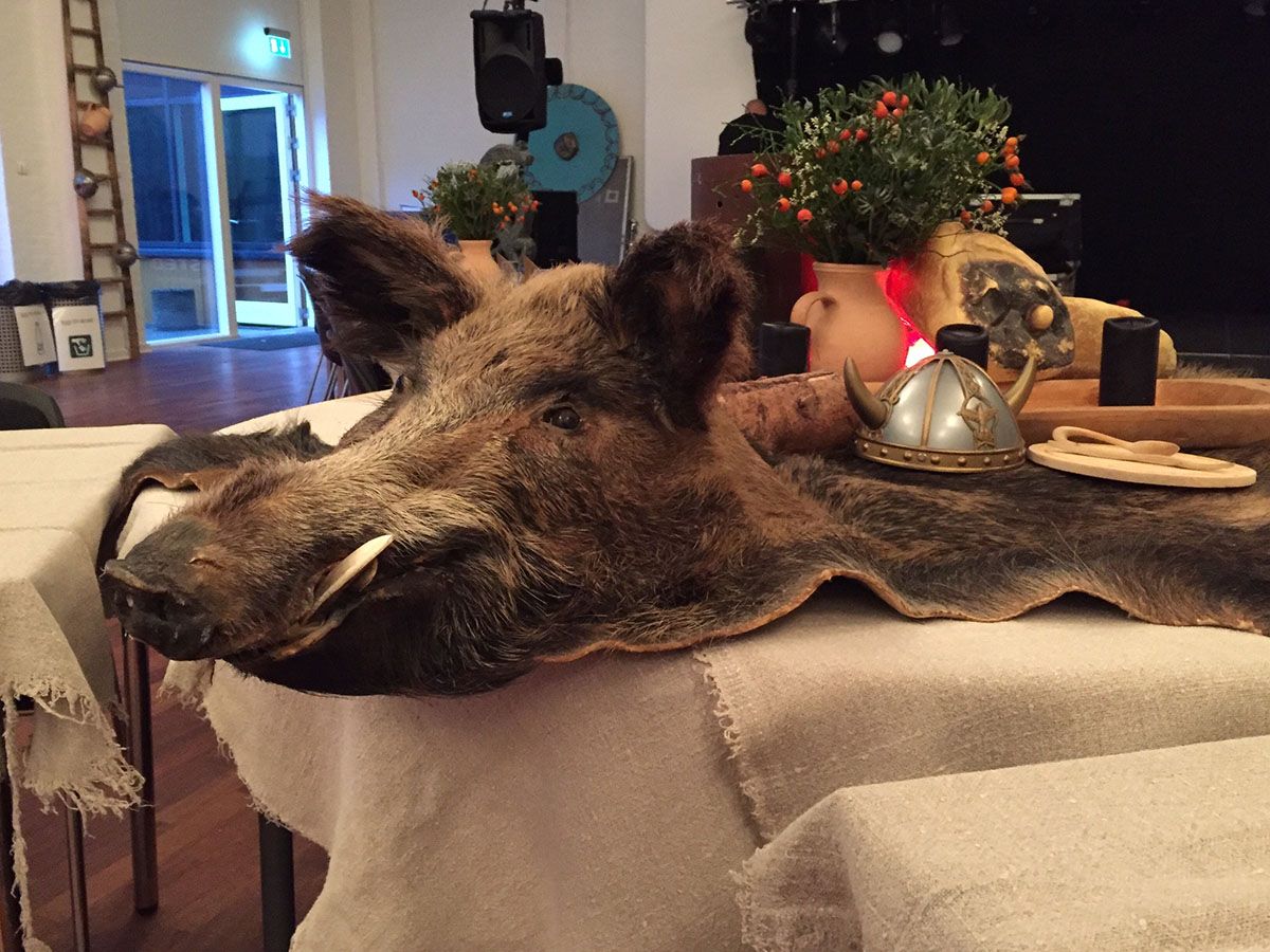 Et vildsvineskind med hoved ligger på bordet. Ovenpå ligger en vikingehjelm og træservice