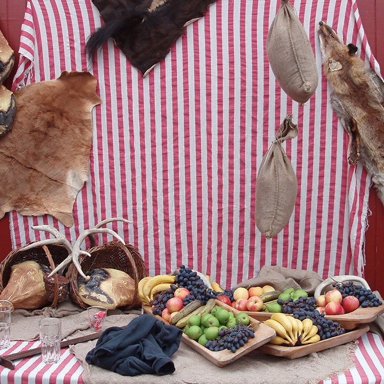 Et bord er pyntet med en rod og hvid stribet dug, hvor der ligger frugter på fade, dyrekøller og gevirer på. På væggen bagved hænger dyreskind og sandsække 