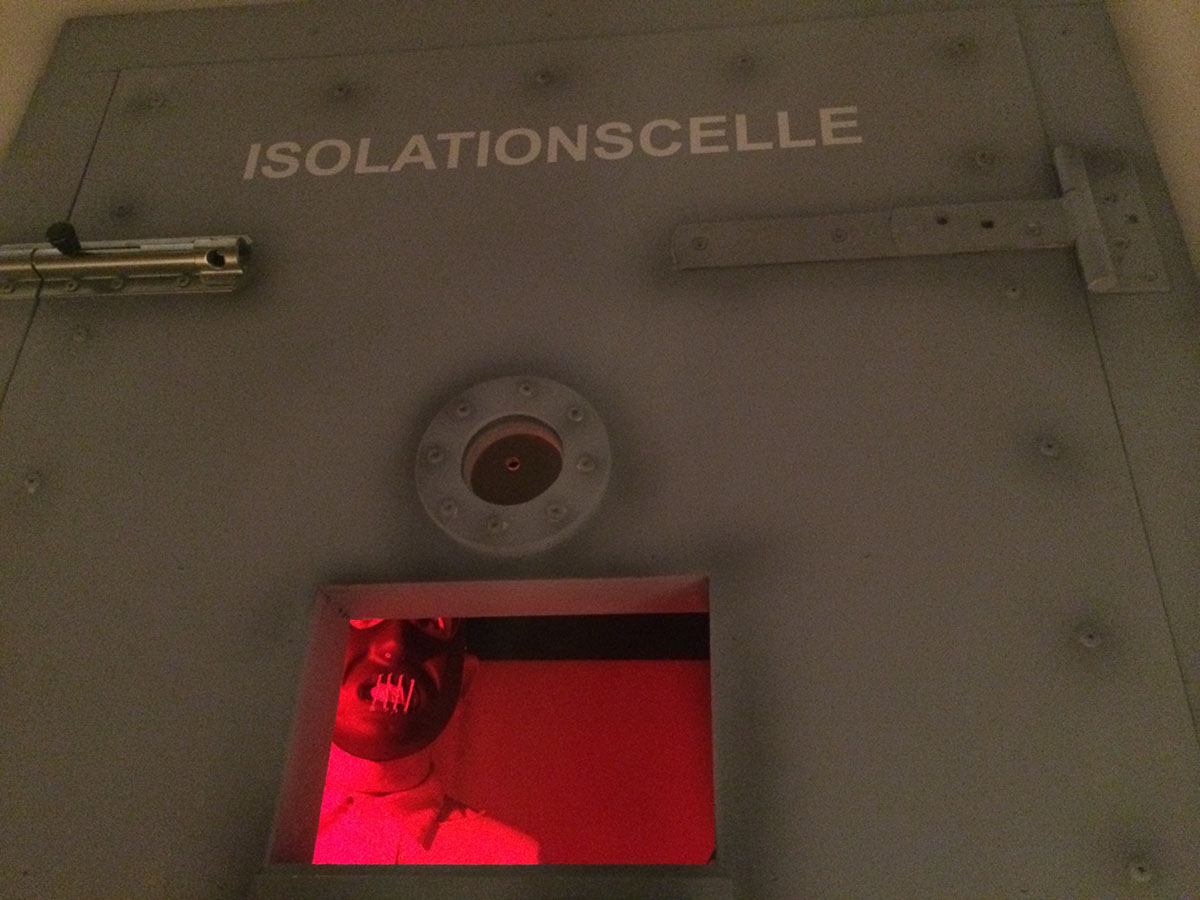 Gæsterne kan kigge ind i en isolationscelle, hvor en mand med en maske står i rødt lys 