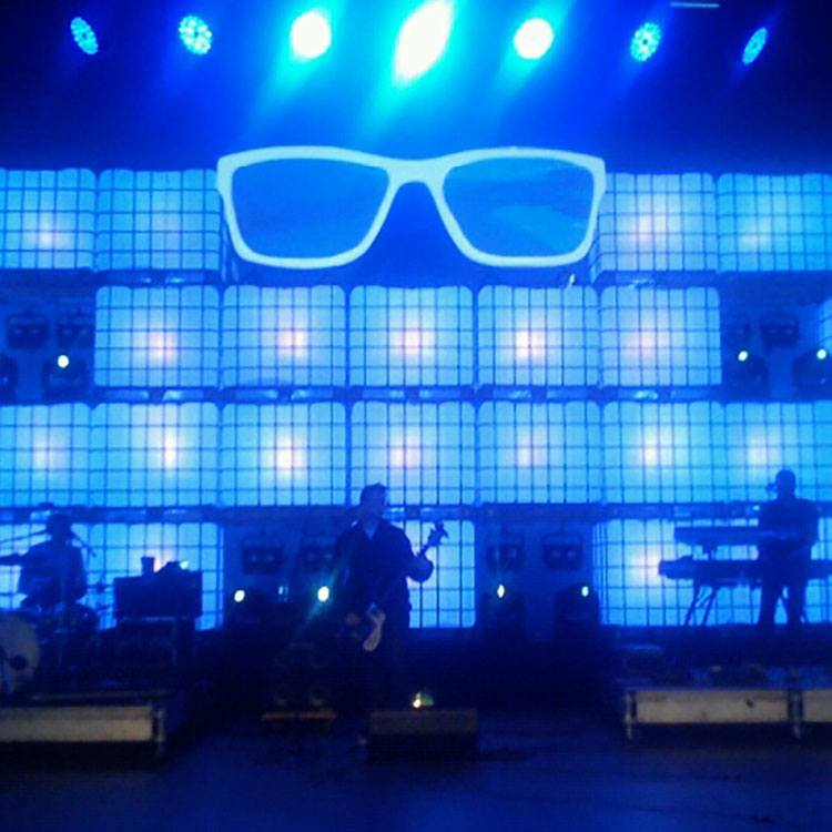 Et band spiller i forgrunden af en masse farvede vandtanke og et par kæmpe briller i blåt lys 