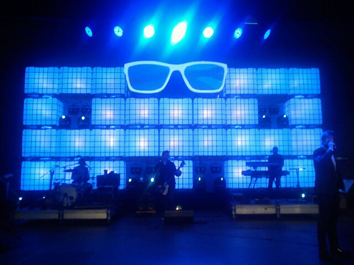 Et band spiller i forgrunden af en masse farvede vandtanke og et par kæmpe briller i blåt lys 