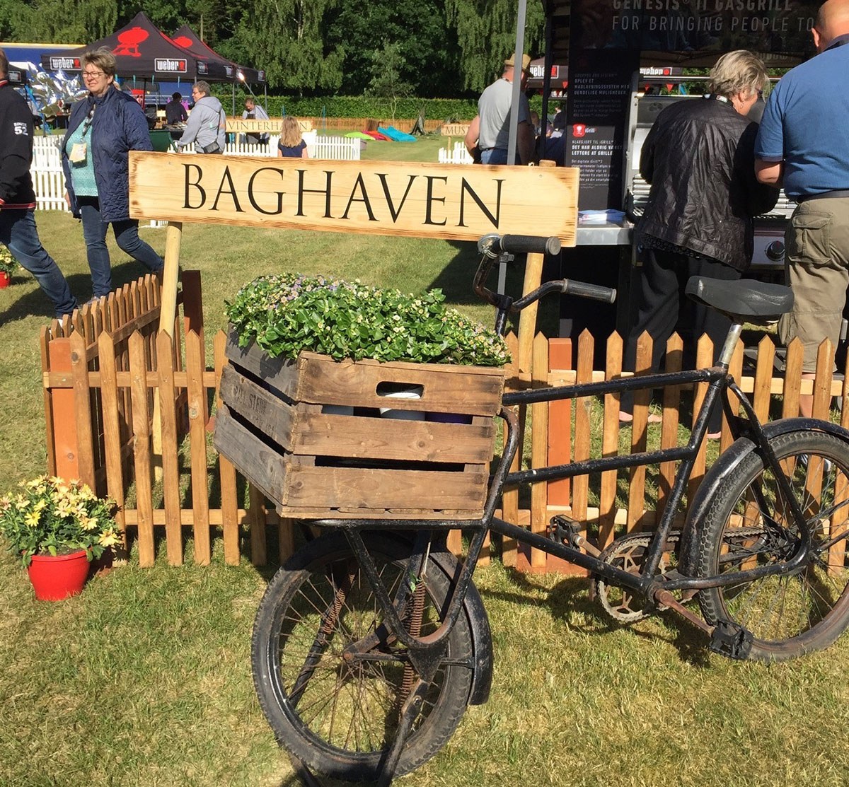 En gammel cykel står med en trækasse fyldt med blomster. Bag ved står et skilt, hvor på der står 'baghaven'
