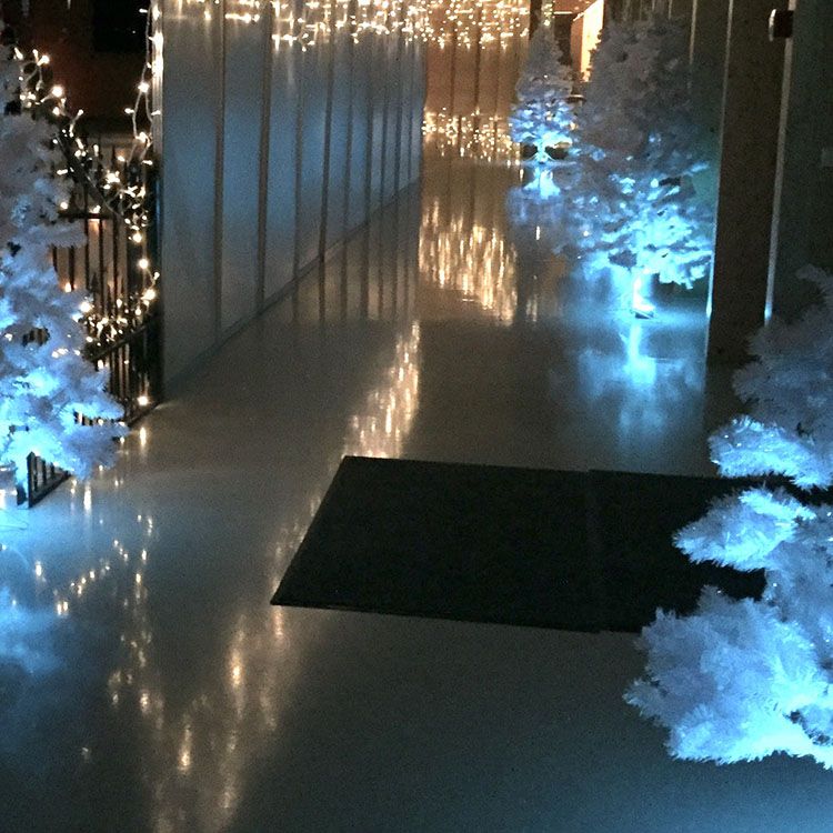 Hvide juletræer med lys på står langs gangen. Langs væggene hænger 
der lyskæder
